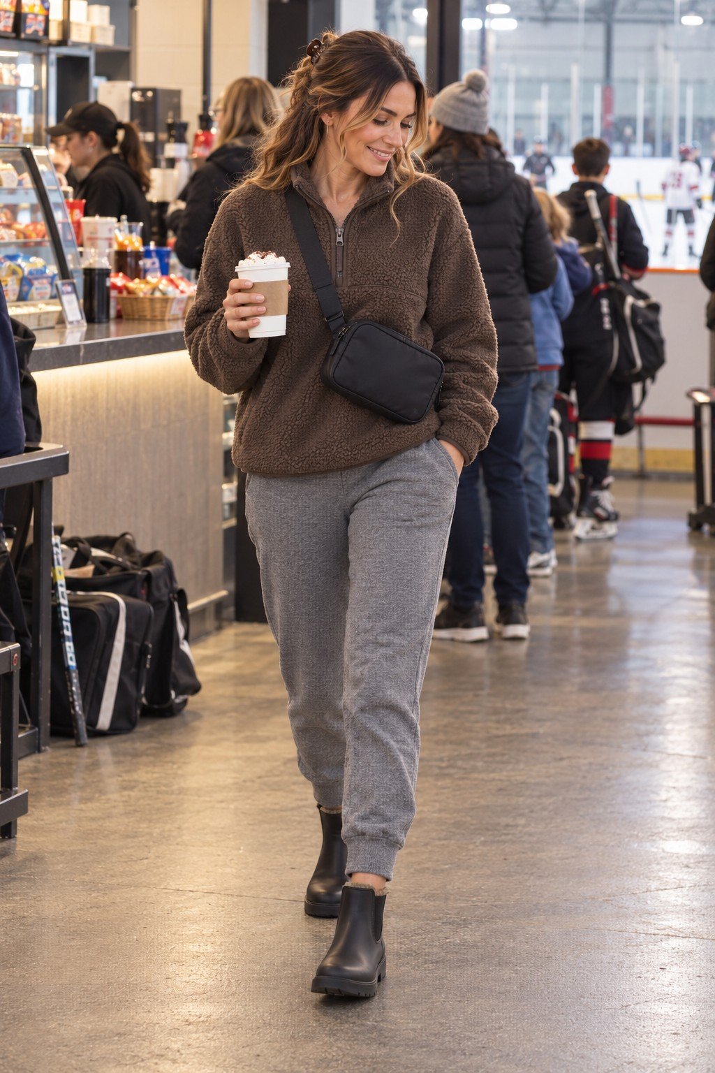 Hockey mom in a brown sherpa quarter zip, gray joggers, chelsea boots and a belt bag at the rink snack bar
